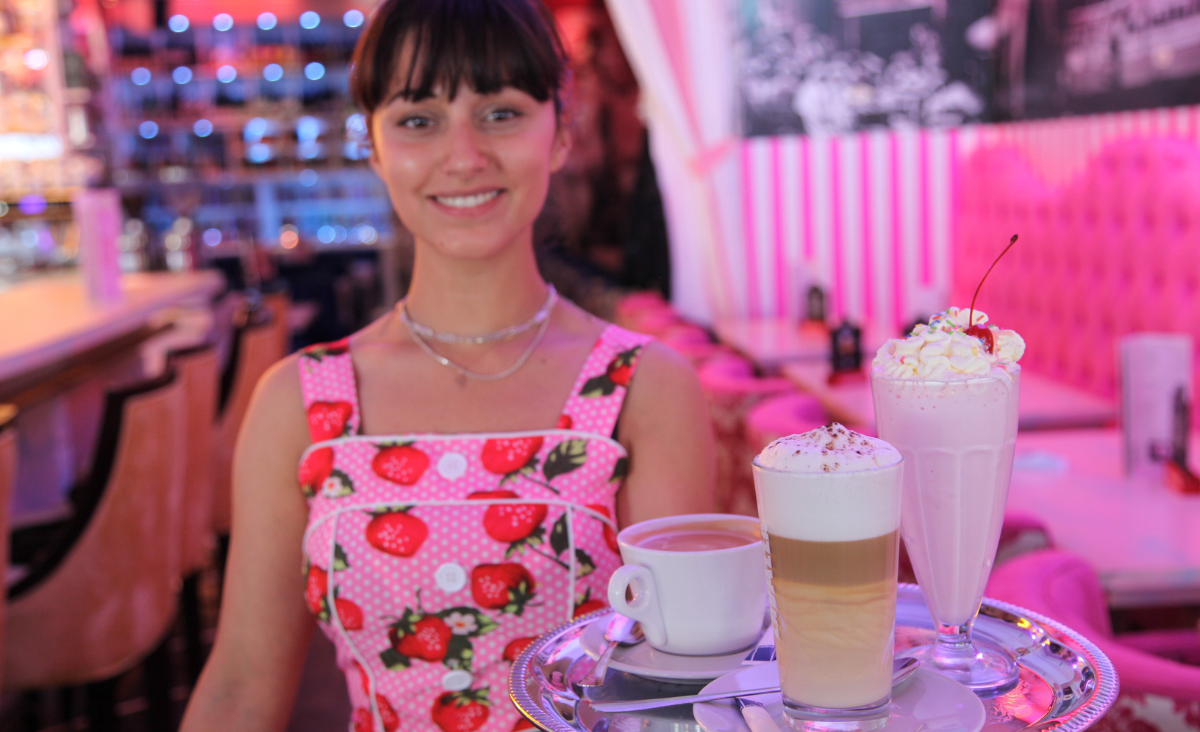 Eine Frau in einem rosa Kleid hält ein Tablett mit zwei Getränken in der Hand.