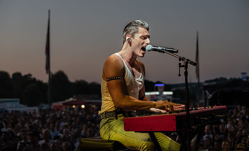 Ein Mann in einem Tanktop singt in ein Mikrofon und spielt dabei Piano.