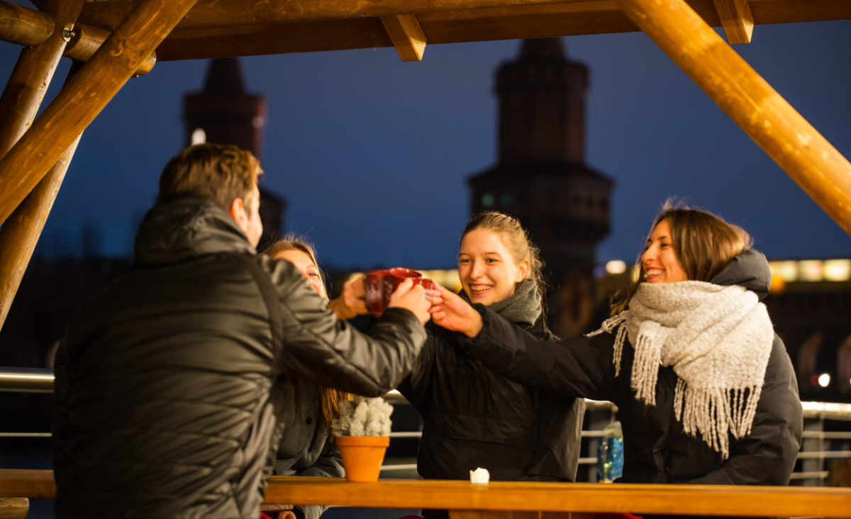 Drei Personen stoßen an einem Tisch an, im Hintergrund ist ein Gebäude zu sehen.