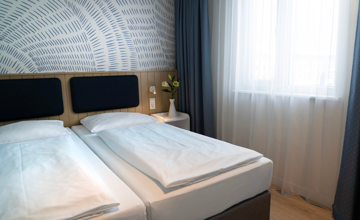 Ein gemütliches Bett mit weißen Bettlaken vor einer blauen, gemusterten Wand.