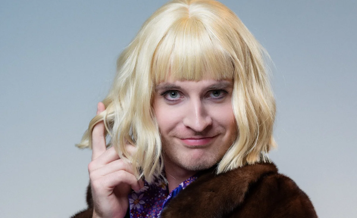 Ein Mann mit blonden Haaren trägt einen Pelzmantel und blickt in die Kamera.
