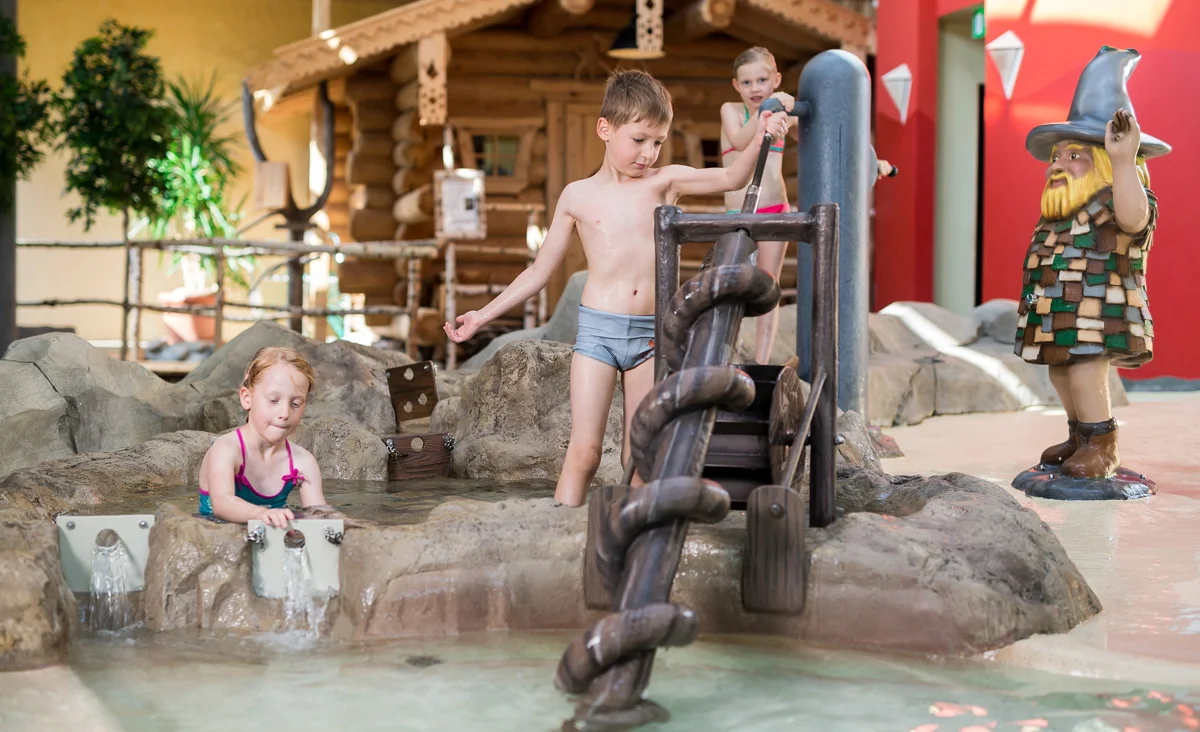 Kinder spielen in einem Wasserbecken mit einer hölzernen Wassermühle und einem kleinen Wasserfall in den Badegärten Eibenstock