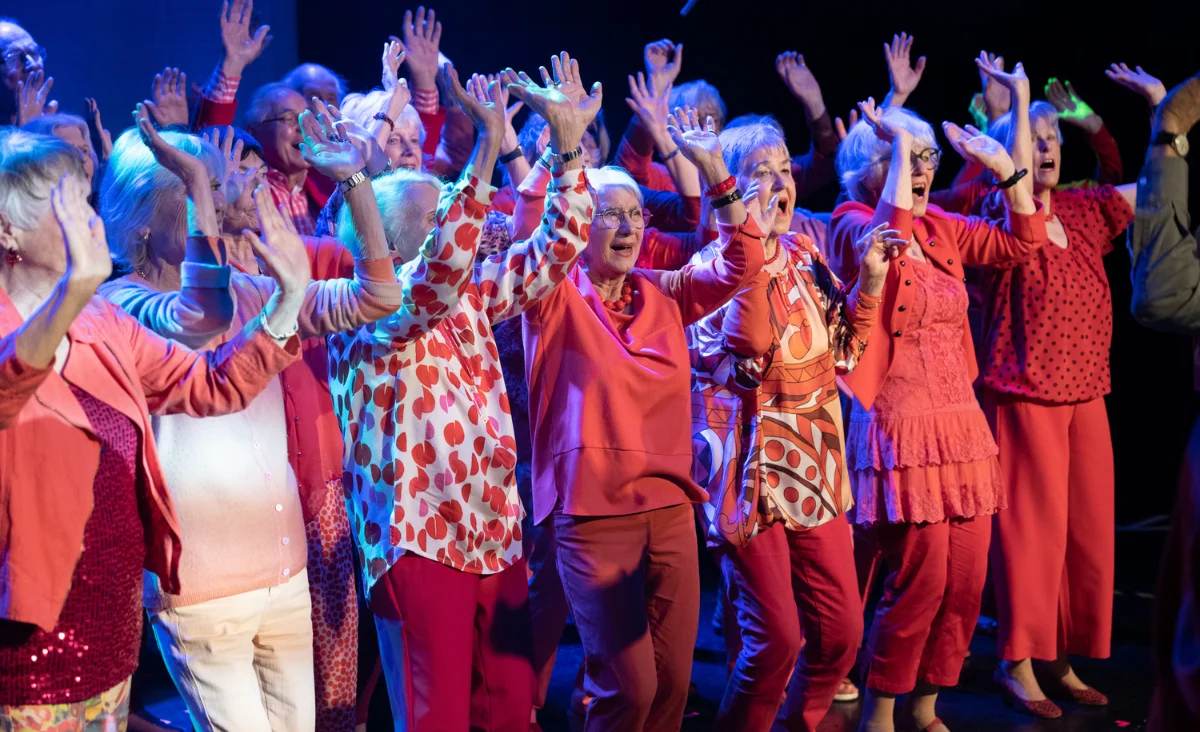 Gruppe Senioren in roten Outfits, eine Frau hebt die Hände, während sie "Heaven can wait" im Chor singen.