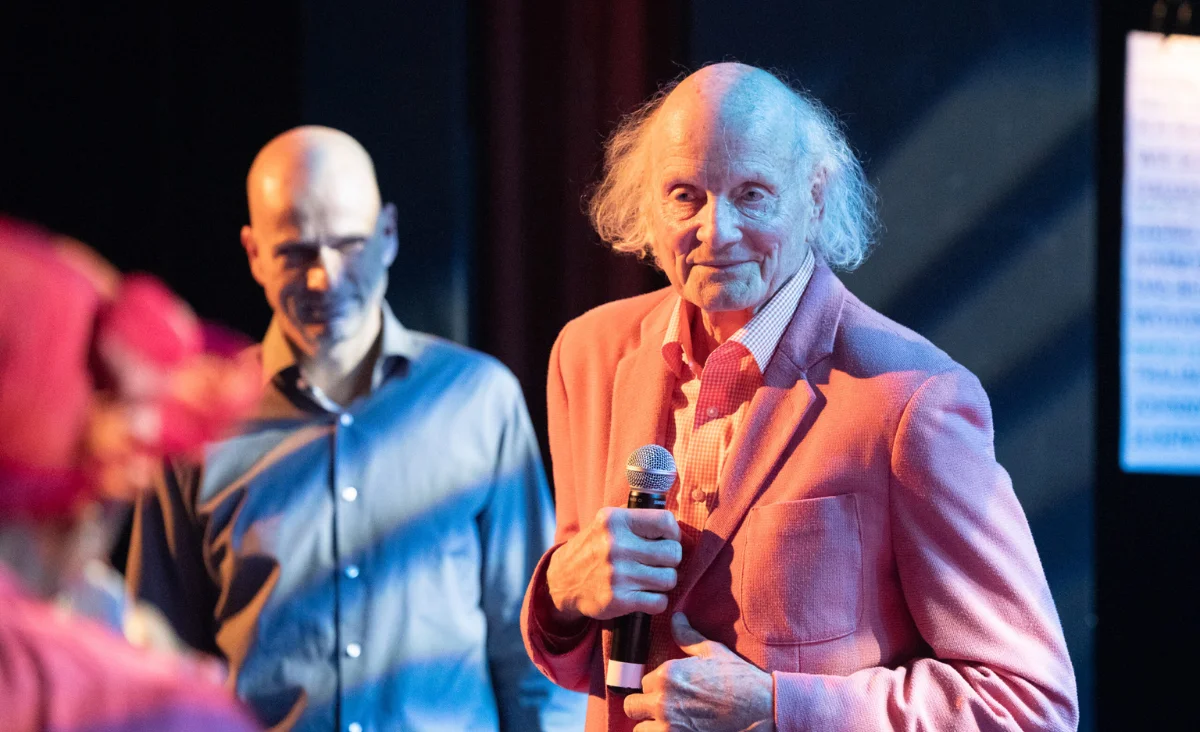Ein Mann im pinken Anzug hält ein Mikrofon und steht vor dem Chor "Heaven can wait".