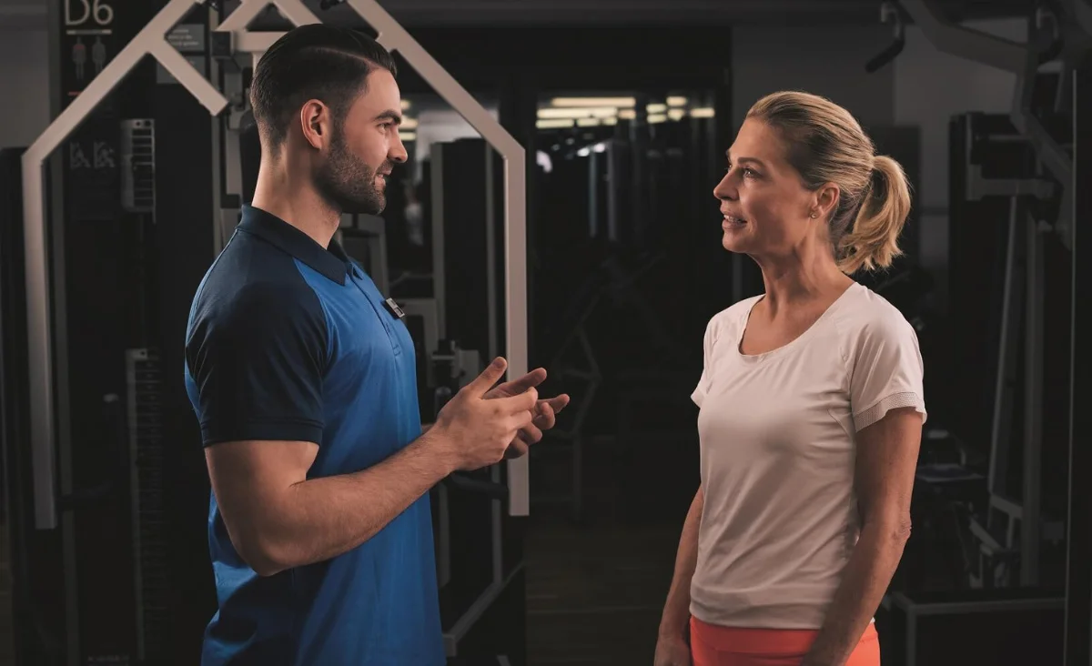 Trainer in blauem Poloshirt spricht mit Frau in weißem T-Shirt im Kieser Studio mit Trainingsgeräten im Hintergrund