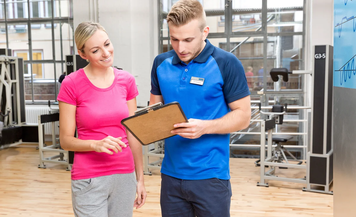 Trainer in blauem Poloshirt spricht mit Frau in pinkem T-Shirt im Kieser Studio mit Trainingsgeräten im Hintergrund