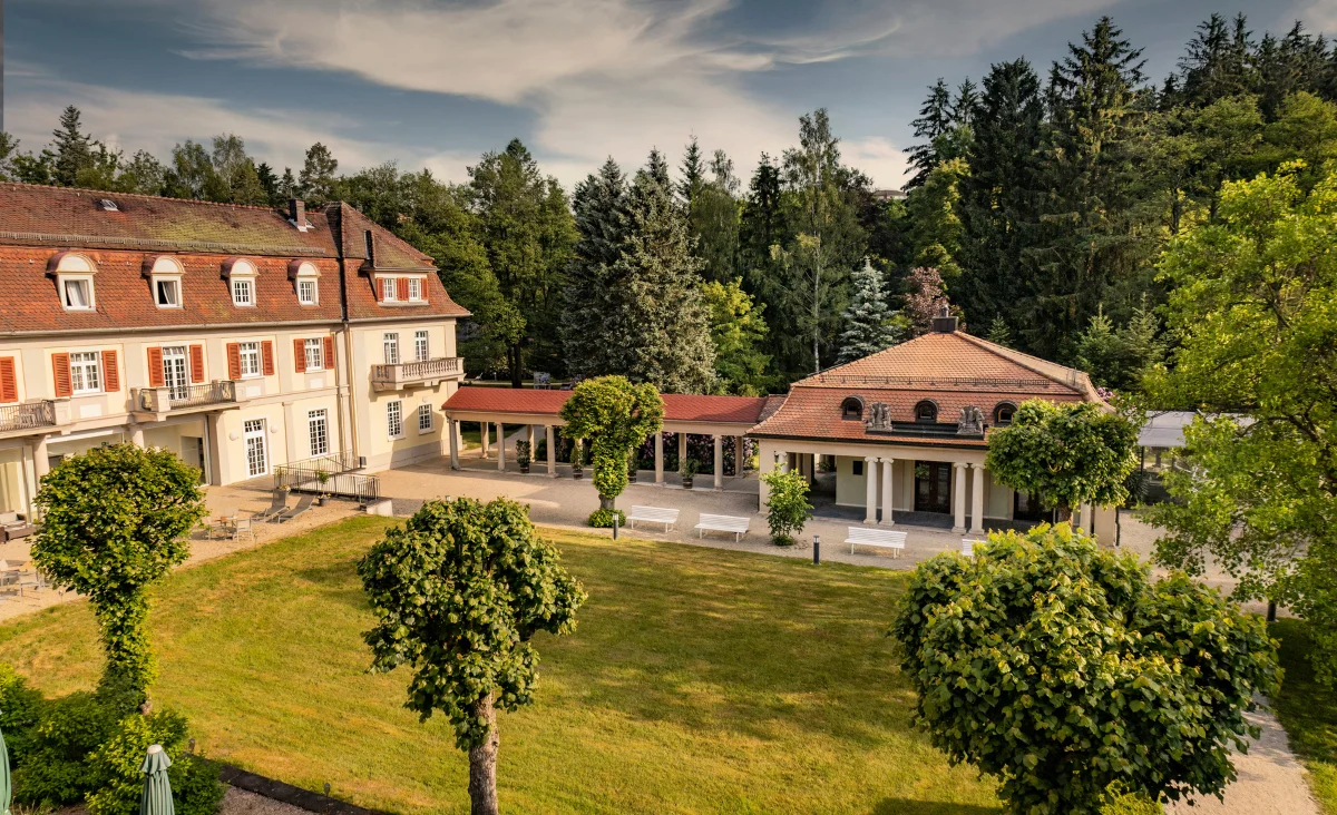 Das Santé Royale Hotel- & Gesundheitsresort Bad Brambach ist von einer gepflegten Gartenanlage mit kurz geschnittenem Rasen und mehreren beschnittenen Bäumen umgeben. Das Hauptgebäude links zeigt eine Fassade mit roten Dachziegeln, weißen Fensterrahmen und mehreren Gauben. Rechts schließt sich ein Nebengebäude mit Säulengang und überdachtem Eingangsbereich an. Im Hintergrund erstreckt sich ein dichter, grüner Mischwald. Die Szene ist von warmem Tageslicht erhellt, der Himmel zeigt einige Wolken.
