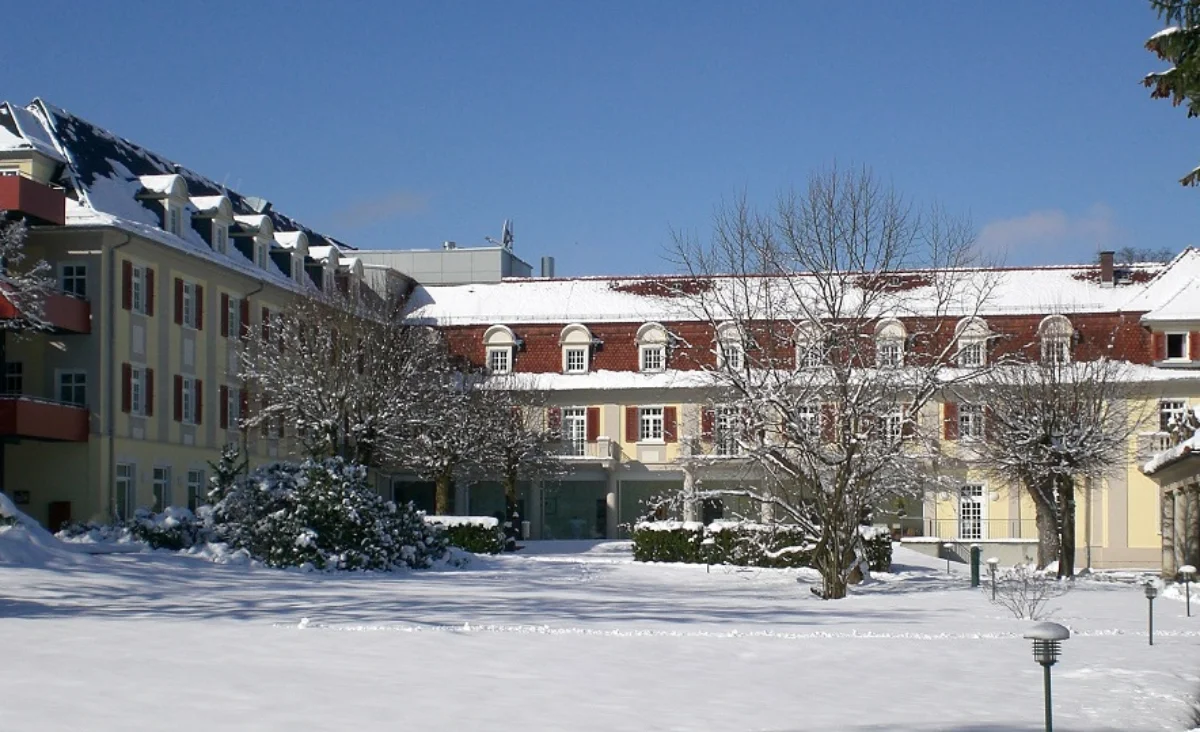 Das Santé Royale Hotel- & Gesundheitsresort Bad Brambach ist von einer geschlossenen Schneedecke umgeben. Die Fassade des mehrstöckigen Gebäudes zeigt helle, gelbe und rote Farbtöne mit weißen Fensterrahmen. Vor dem Gebäude stehen mehrere laublose Bäume, deren Äste mit Schnee bedeckt sind. Der Himmel ist wolkenlos und blau. Im Vordergrund erstreckt sich eine verschneite Rasenfläche, auf der vereinzelt Sträucher und ein kleiner Laternenmast zu sehen sind.