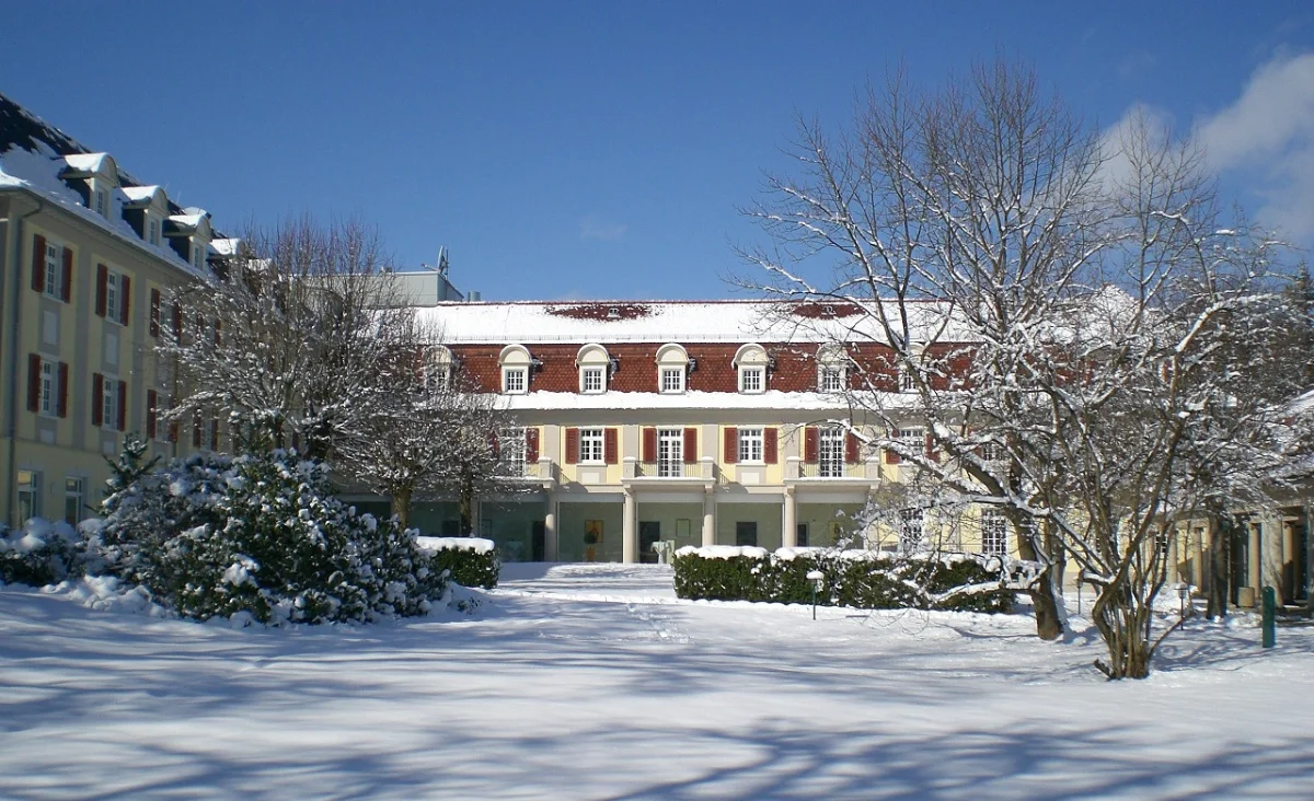 Das Santé Royale Hotel- & Gesundheitsresort Bad Brambach ist von einer geschlossenen Schneedecke umgeben. Die Fassade des mehrstöckigen Gebäudes zeigt helle, gelbe und rote Farbtöne mit weißen Fensterrahmen. Vor dem Gebäude stehen mehrere laublose Bäume, deren Äste mit Schnee bedeckt sind. Der Himmel ist wolkenlos und blau. Im Vordergrund erstreckt sich eine verschneite Rasenfläche, auf der vereinzelt Sträucher und ein kleiner Laternenmast zu sehen sind.