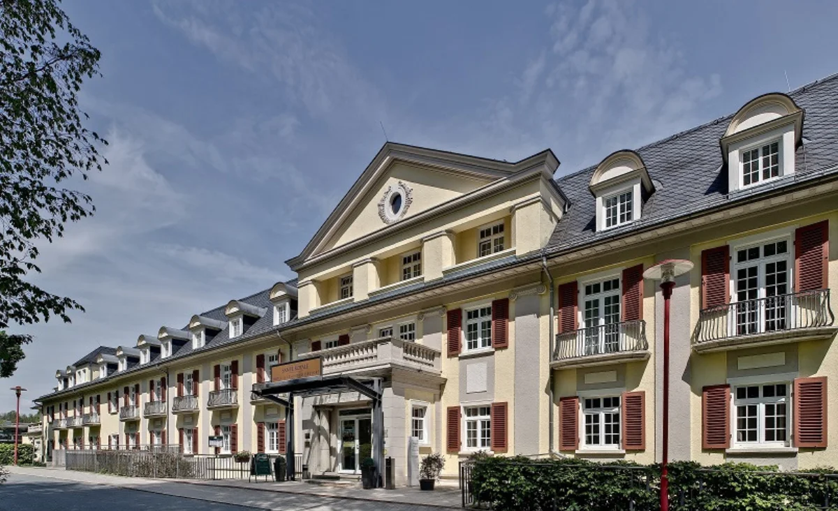 Das Santé Royale Hotel- & Gesundheitsresort Bad Brambach zeigt eine langgestreckte, dreigeschossige Fassade mit gelbem Anstrich und weißen Fensterrahmen. Die Fensterläden sind in einem dunklen Rotton gehalten. Im mittleren Bereich befindet sich ein überdachter Eingangsbereich mit Vordach und Säulen. Das Dach ist dunkel gedeckt und weist mehrere Gauben auf. Vor dem Gebäude stehen niedrige Hecken, ein Gehweg und eine Straßenlaterne. Der Himmel ist leicht bewölkt.