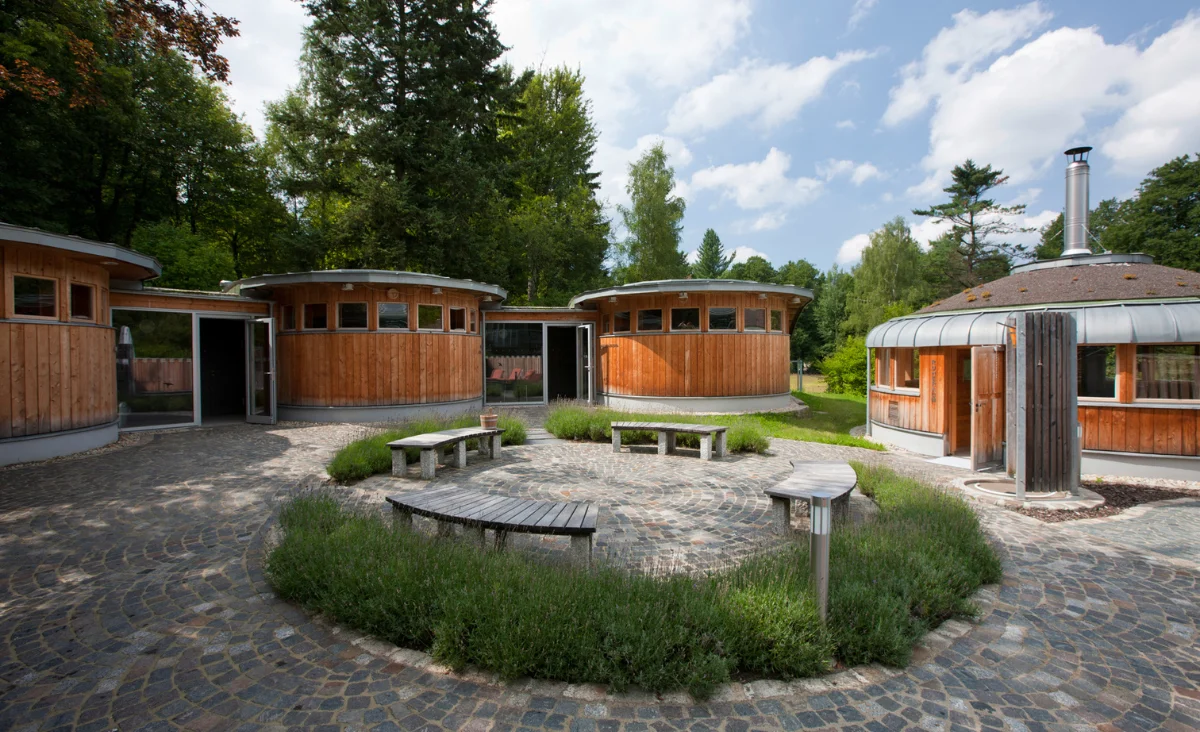 Holzverkleidete Rundbauten mit flachen Dächern stehen in einem gepflasterten Innenhof, der von niedrigen Lavendelbeeten eingefasst ist. Im Zentrum des Hofs befinden sich mehrere geschwungene Holzbänke. Die Gebäude gehören zum Santé Royale Hotel- & Gesundheitsresort Bad Brambach. Im Hintergrund sind hohe Bäume und blauer Himmel mit einigen Wolken zu sehen.