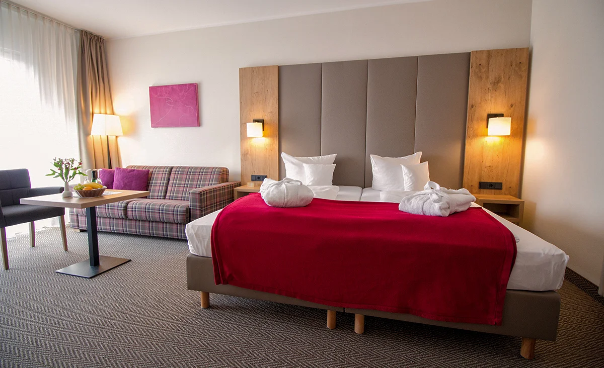Hotelzimmer mit Doppelbett, gepolstertem Kopfteil, rot-weiß kariertem Sofa, grauem Sessel, Holzmöbeln und pinkfarbenem Wandbild in Hotel Wolkenstein