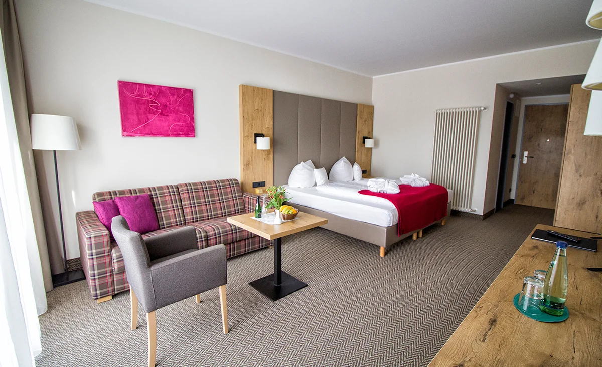 Hotelzimmer mit Doppelbett, gepolstertem Kopfteil, rot-weiß kariertem Sofa, grauem Sessel, Holzmöbeln und pinkfarbenem Wandbild in Hotel Wolkenstein