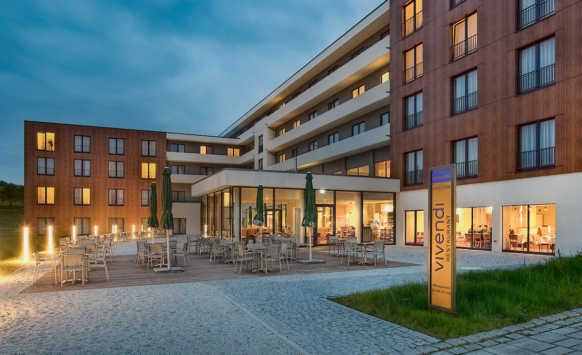 Moderner mehrstöckiger Hotelbau mit Terrasse, Tischen, Stühlen und geschlossenen Sonnenschirmen bei Dämmerung