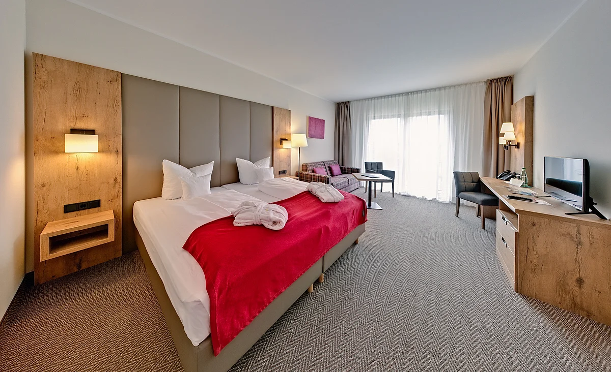 Hotelzimmer mit Doppelbett, gepolstertem Kopfteil, rot-weiß kariertem Sofa, grauem Sessel, Holzmöbeln und pinkfarbenem Wandbild in Hotel Wolkenstein