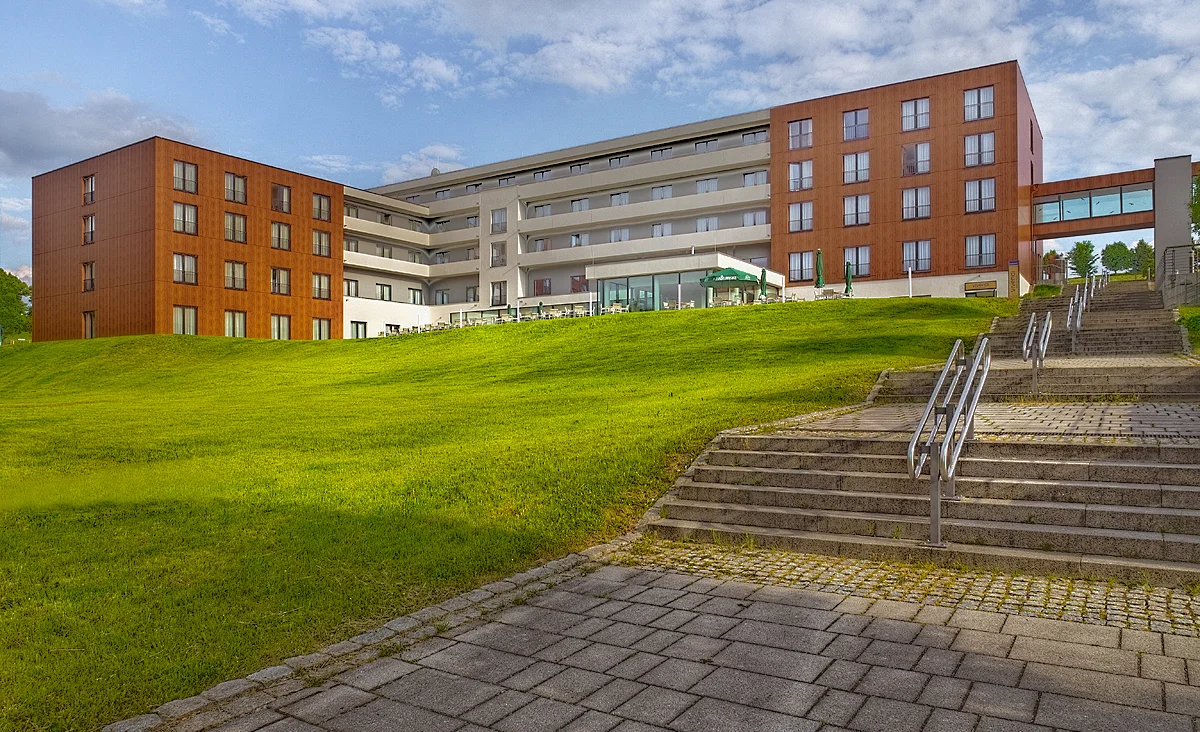 Grünfläche mit quadratischem Glasobjekt vor modernem Gebäude mit Schriftzug 'SILBER-THERME' und überdachtem Verbindungssteg