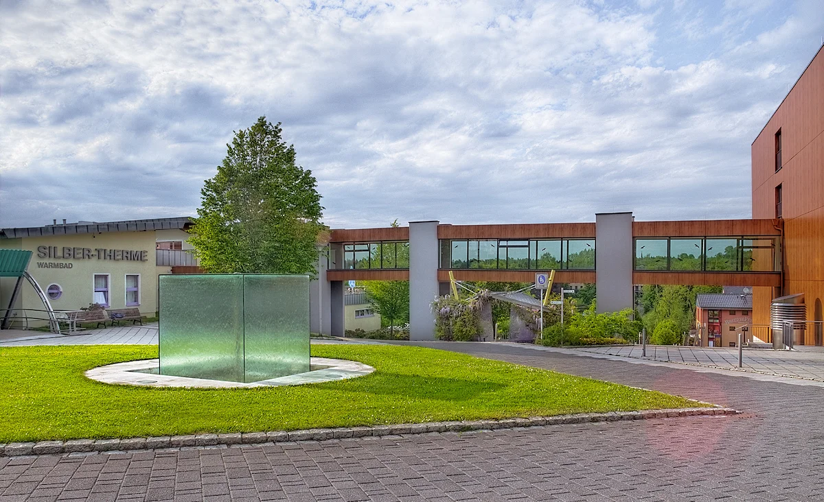 Grünfläche mit quadratischem Glasobjekt vor modernem Gebäude mit Schriftzug 'SILBER-THERME' und überdachtem Verbindungssteg