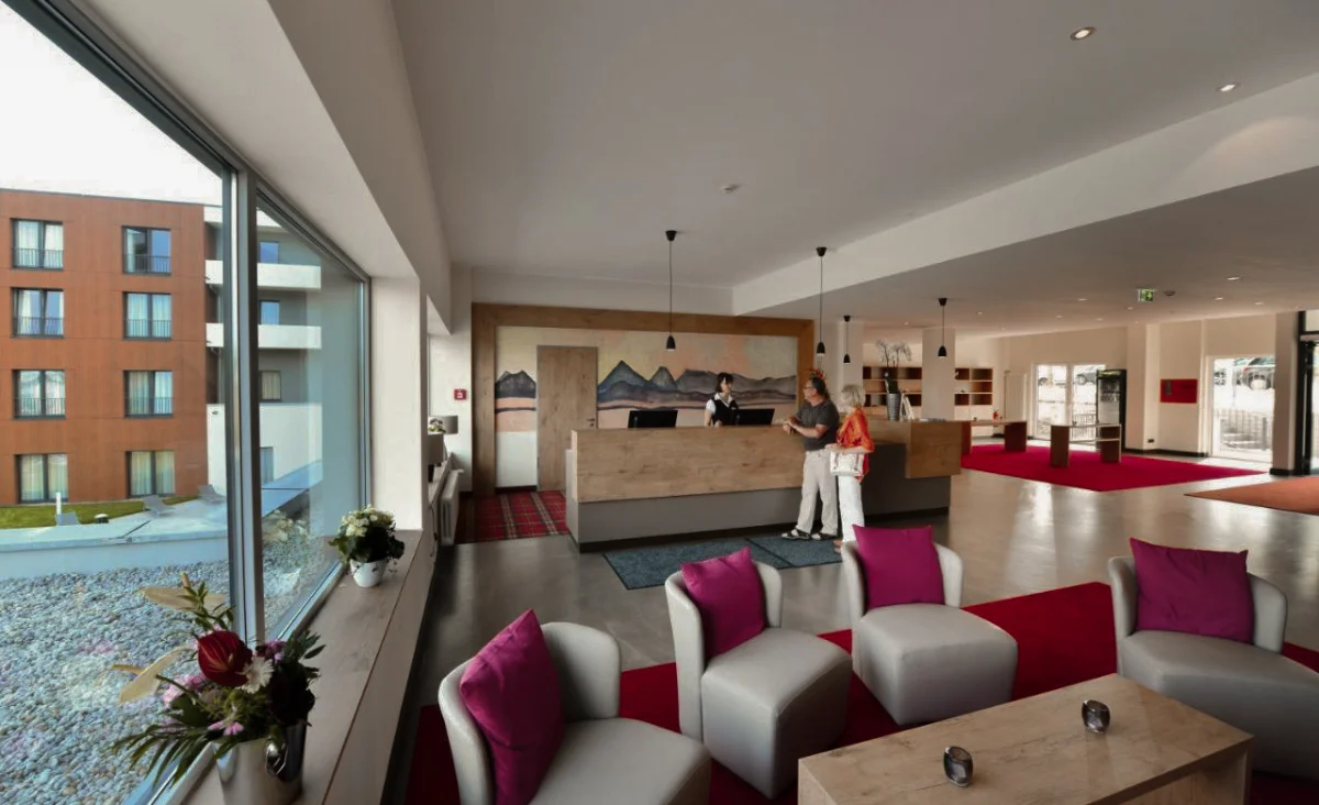 Hotel-Lobby mit moderner Holzrezeption, zwei Personen am Empfang, Sitzgruppe mit grauen Sesseln und roten Kissen sowie großem Fenster mit Blick auf ein benachbartes Gebäude