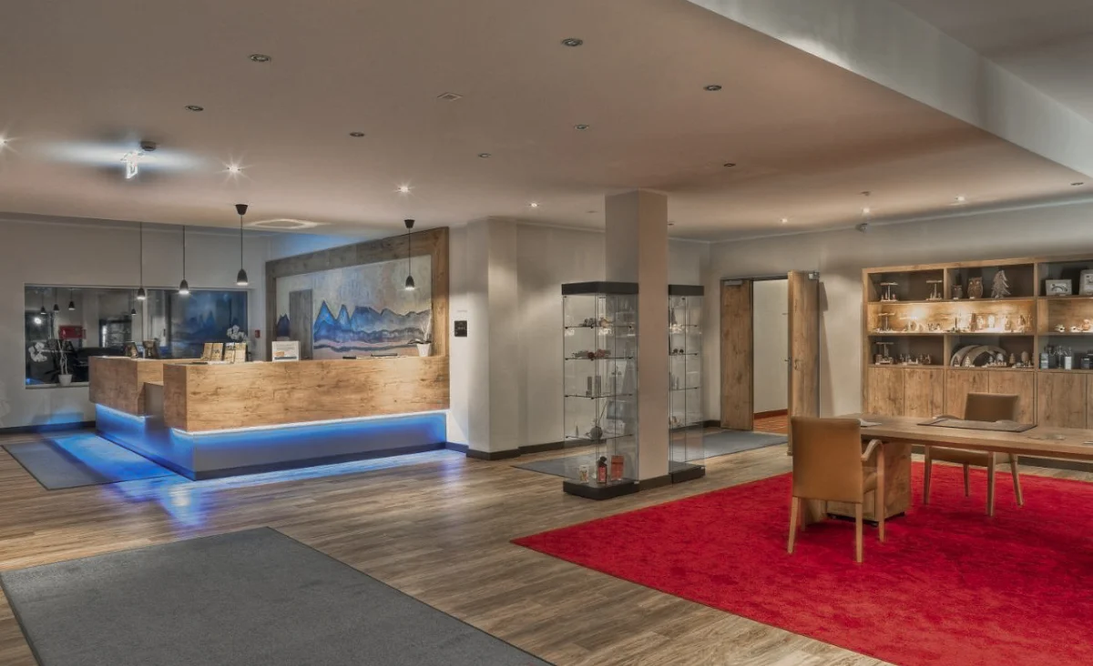 Moderner Hotel-Empfangsbereich mit beleuchtetem Holztresen, rotem Teppich, Sitzmöbeln und Vitrinen im Hotel Wolkenstein