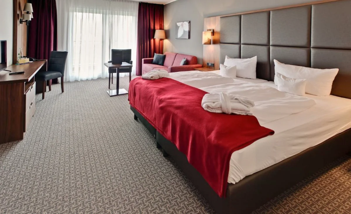 Hotelzimmer mit Doppelbett, gepolstertem Kopfteil, rot-weiß kariertem Sofa, grauem Sessel, Holzmöbeln und pinkfarbenem Wandbild in Hotel Wolkenstein