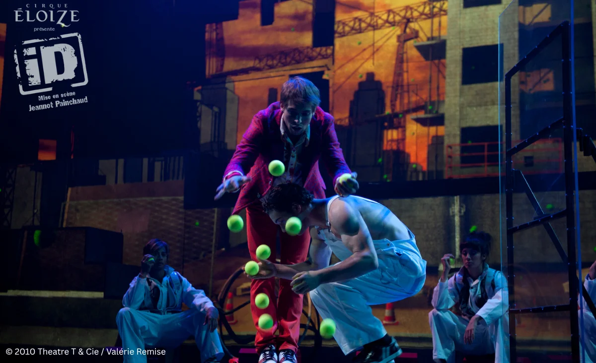 Cirque Éloize: Ein Mann jongliert Tennisbälle, während er auf einem Stuhl balanciert.