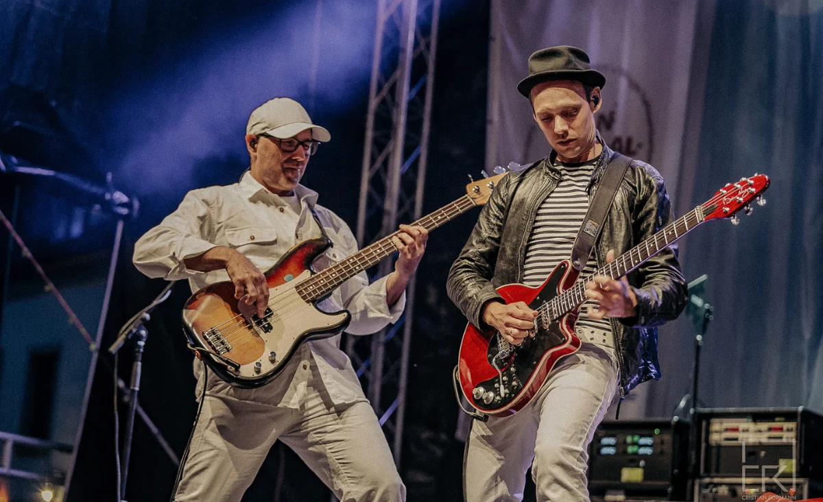 Zwei Männer in weißen Outfits spielen Gitarre auf einer Bühne.