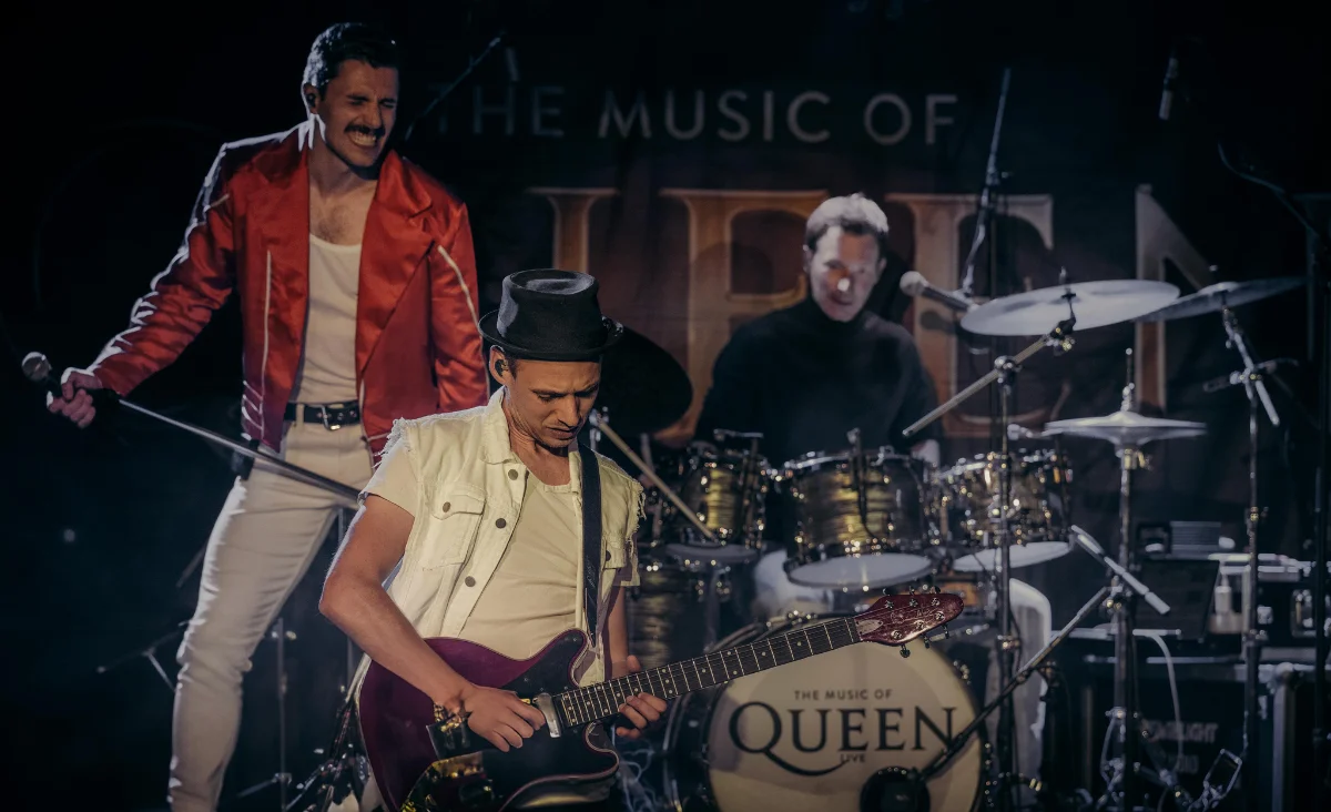Die Band Queen spielt live auf einem Musikfestival, umgeben von begeisterten Fans und bunten Lichtern.