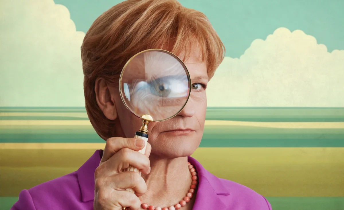 Auf der Bühne von "Miss Merkel in der Komödie am Kurfürstendamm" betrachtet eine Frau etwas durch eine Lupe.