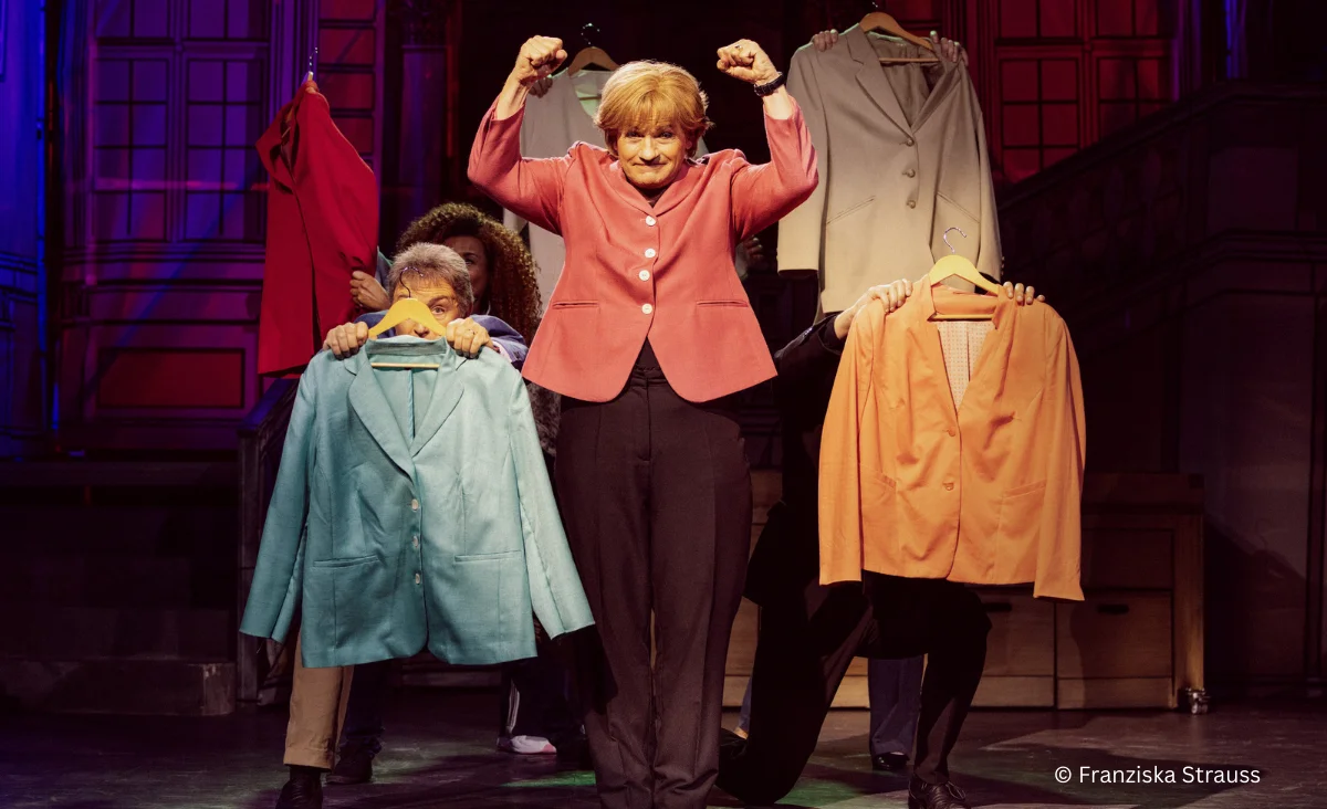 Auf der Bühne präsentiert eine Frau verschiedene Kleidungsstücke in der Komödie "Miss Merkel – Mord in der Uckermark".