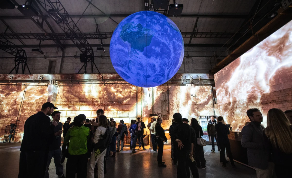 Große blaue Erdkugel schwebt in einer dunklen Ausstellungshalle mit Projektionen an den Wänden und mehreren Menschen, die sich bei der immersiven Ausstellung Origins – Uprung des Lebens in Berlin unterhalten.