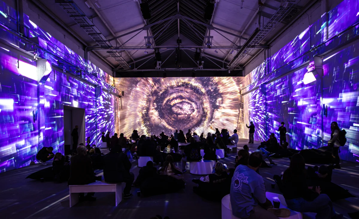 Große Gruppe sitzender und stehender Menschen in einem dunklen Ausstellungsraum mit immersiven, farbigen Projektionen an den Wänden, die ein spiralförmiges Muster an der Stirnwand zeigen