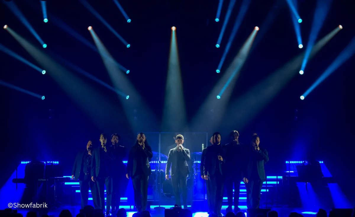 Sieben Sänger in dunkler Kleidung stehen auf einer Bühne mit blauem Bühnenlicht und Scheinwerfern im Hintergrund: The 12 Tenors Konzert in Berlin