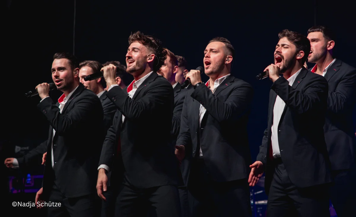 Gruppe von Sänger in schwarzen Anzügen mit weißen Hemden, die auf einer Bühne singen und Mikrofone halten
