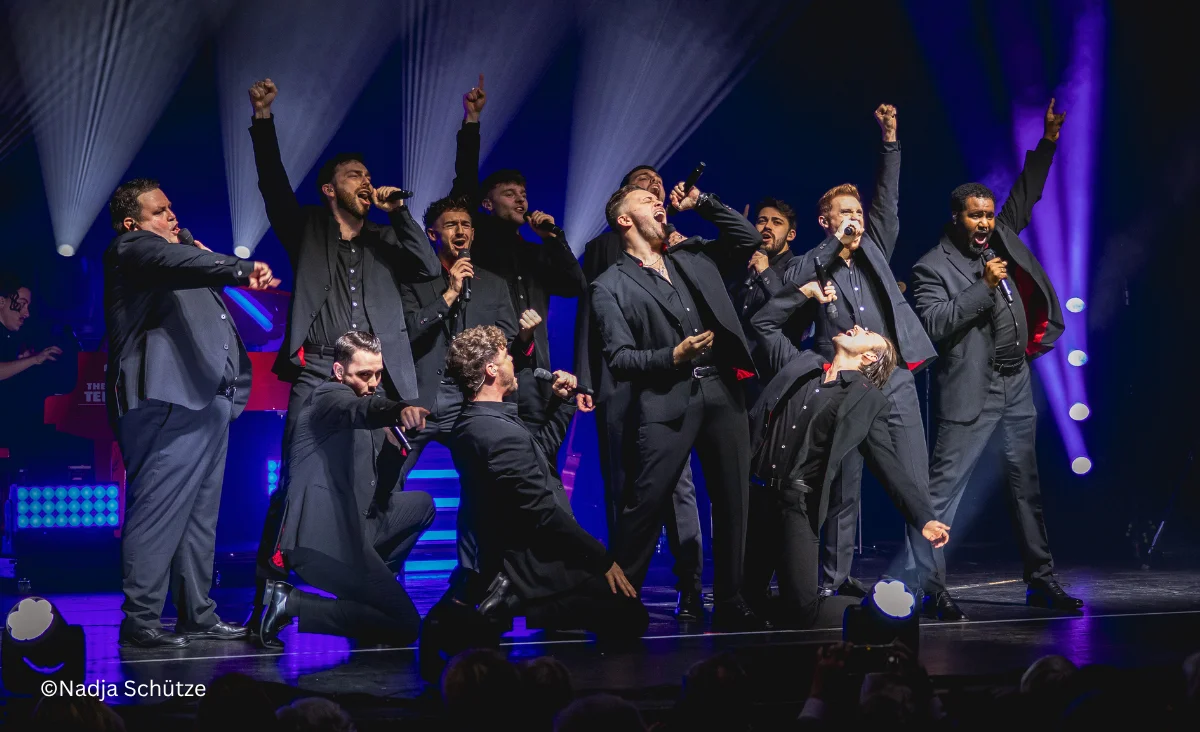 Mehrere Sänger in schwarzen Anzügen auf einer Bühne in dynamischen Posen mit erhobenen Armen und Mikrofonen: The 12 Tenors Konzert Berlin