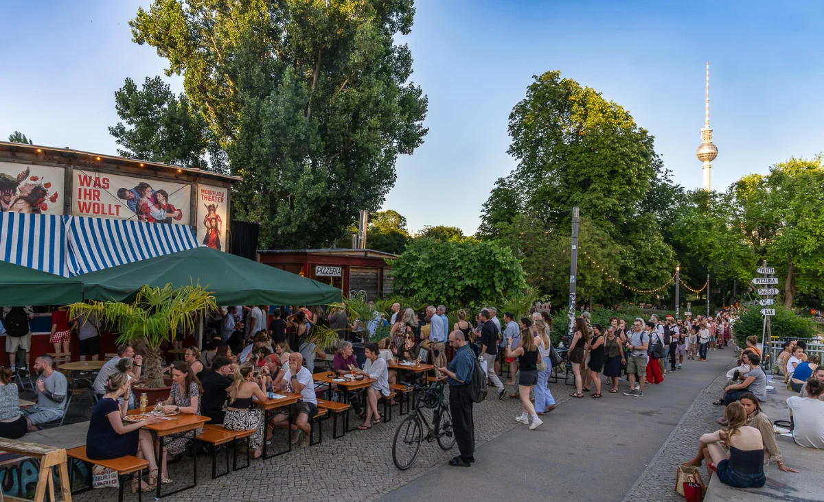 Menschen sitzen und stehen an Tischen unter einem grünen Sonnenschirm vor dem Monbijou Theater Berlin mit Bäumen und dem Fernsehturm im Hintergrund.