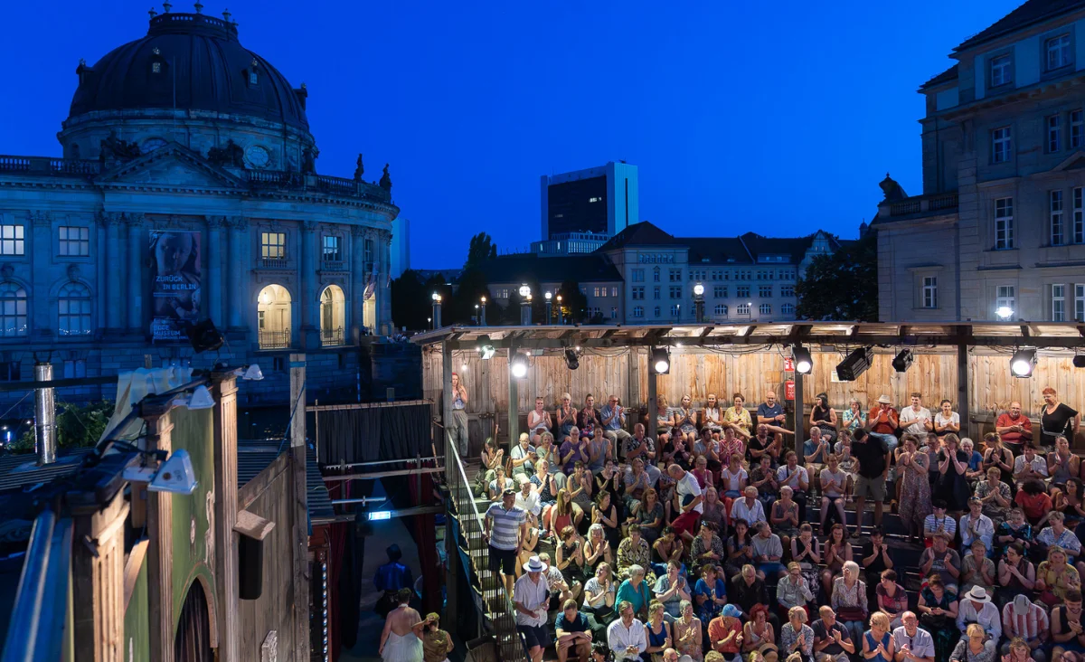 Publikum sitzt bei Abendvorstellung im Monbijou Theater Berlin vor beleuchtetem Bühnenaufbau mit Kuppelgebäude im Hintergrund