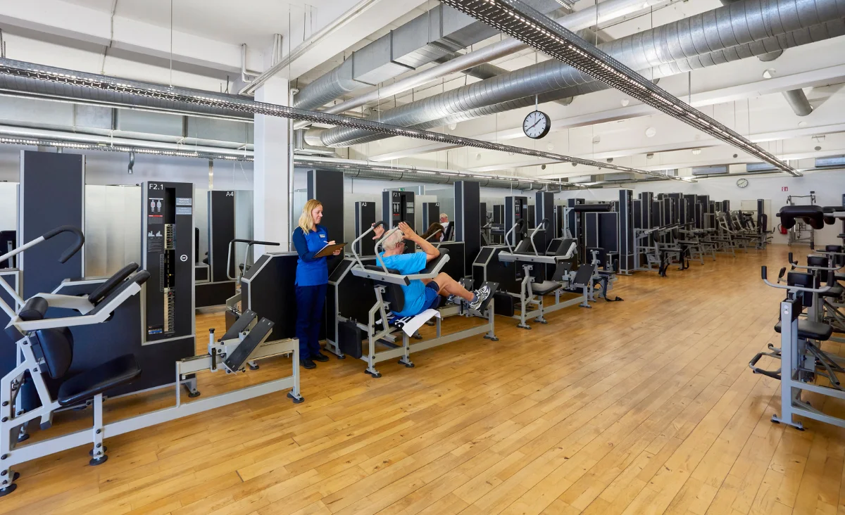 Großer heller Trainingsraum mit mehreren Kieser Training Kraftgeräten, eine Mann in blauer Sportkleidung sitzt an einem Gerät, eine Trainerin mit Tablet steht daneben