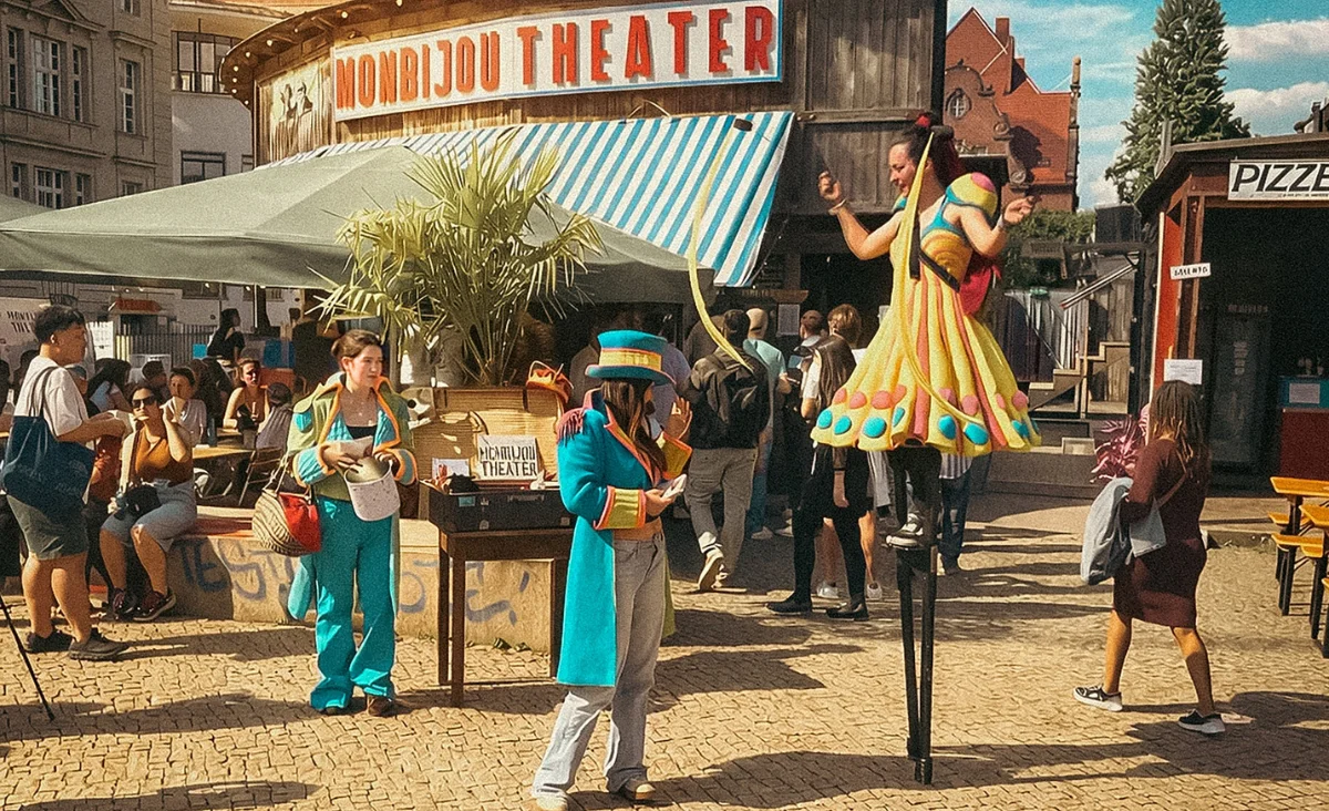 Straßenszene vor dem Monbijou Theater Berlin mit zwei Personen in bunten Kostümen, eine auf Stelzen, umgeben von Passanten und einem Stand mit Theaterplakaten.