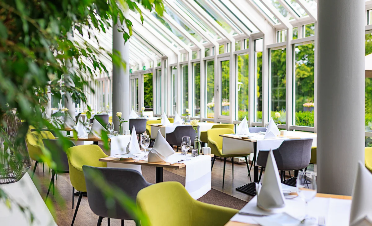 Hell und modern eingerichteter Wintergartenbereich eines Wellnesshotels mit weißen Tischen, grün-grauen Stühlen und großen Glasfenstern mit Blick ins Grüne