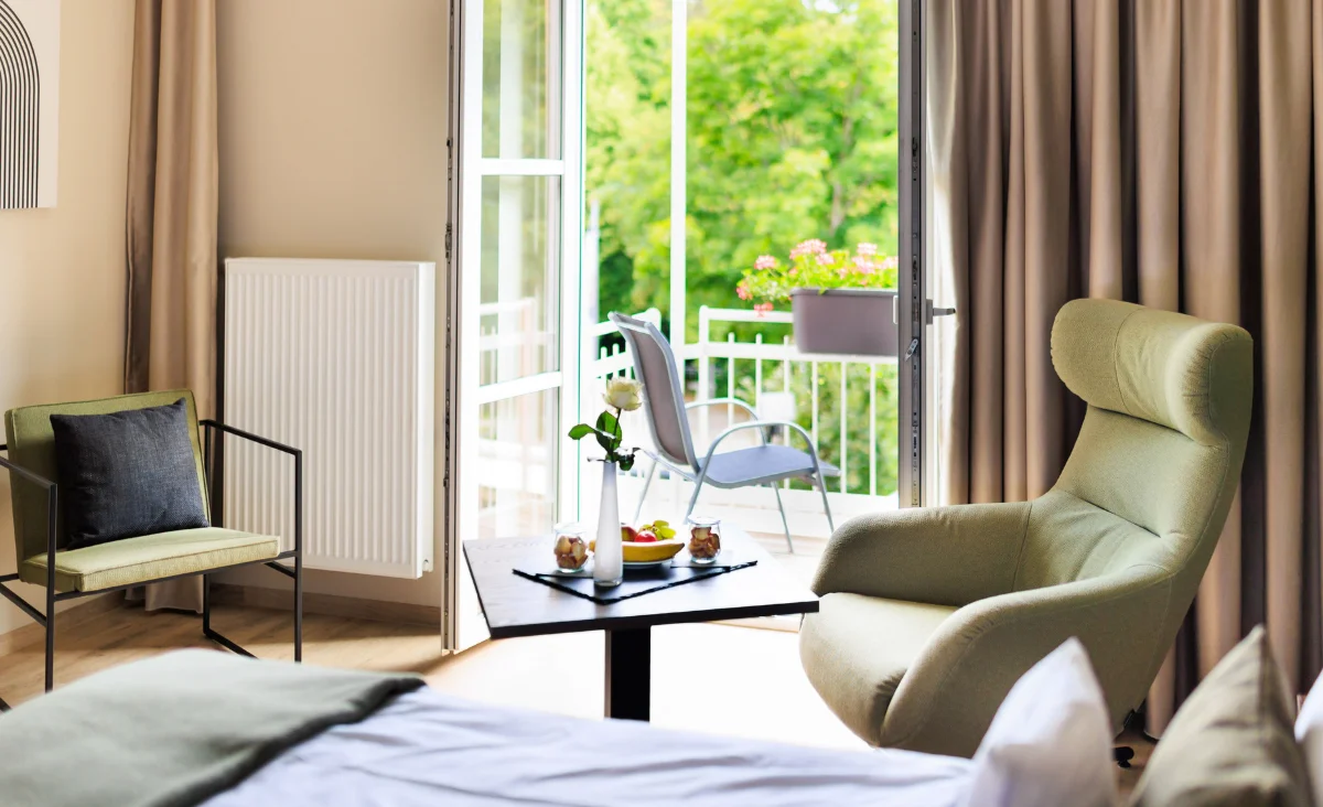 Hotelzimmer mit Sessel, Stuhl, Tisch mit Obst und Getränken vor offenem Balkon mit Blick auf grüne Bäume