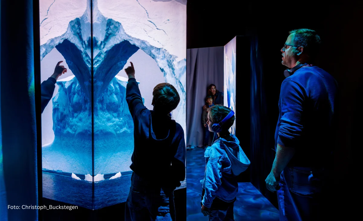 Drei Personen betrachten in der Immersiven Ausstellung „Polar Experience“ eine große, beleuchtete Eisbergprojektion