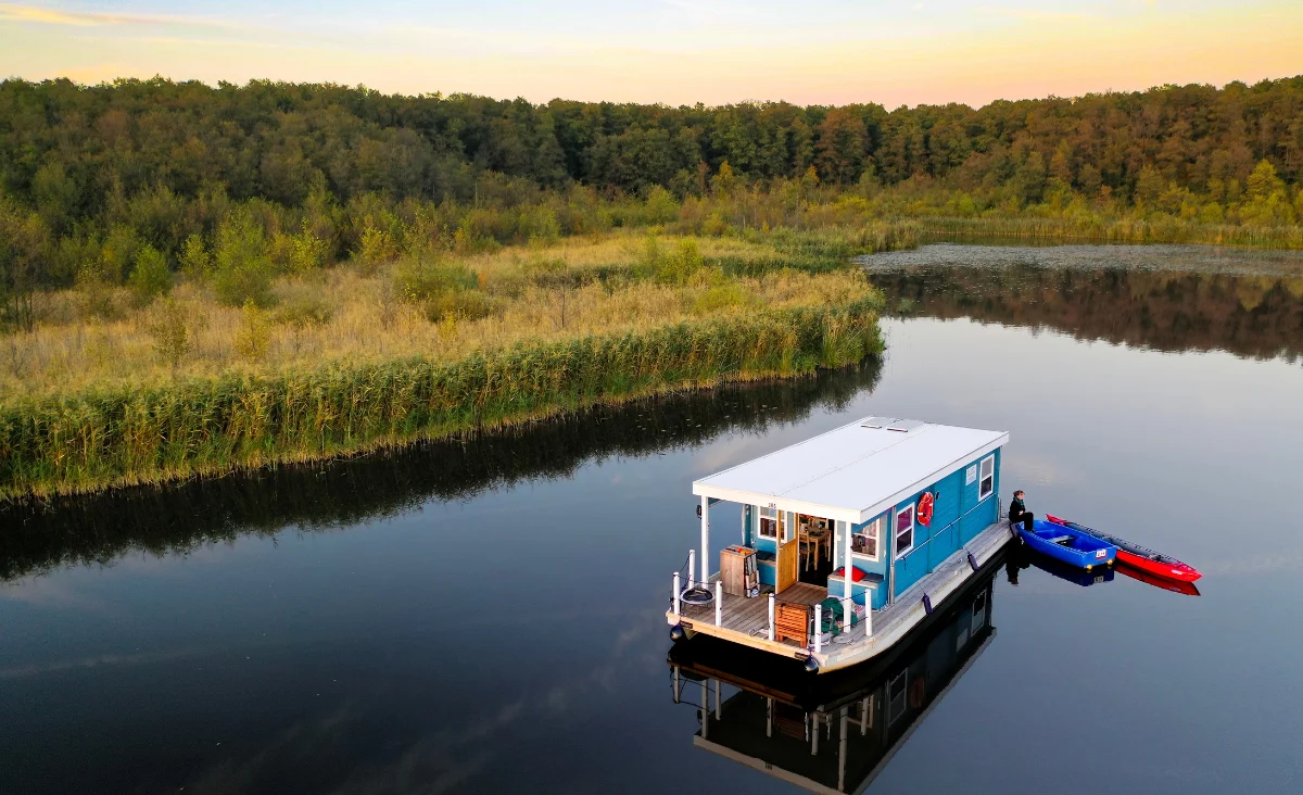 Hausboot fährt auf einem ruhigen Fluss umgeben von grünen Bäumen und mehreren angelegten Booten