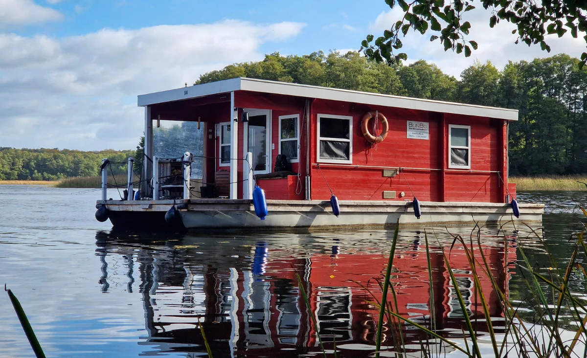 Hausboot mit rechteckigem Aufbau und mehreren Fenstern fährt auf ruhigem See vor bewaldetem Ufer unter klarem Himmel