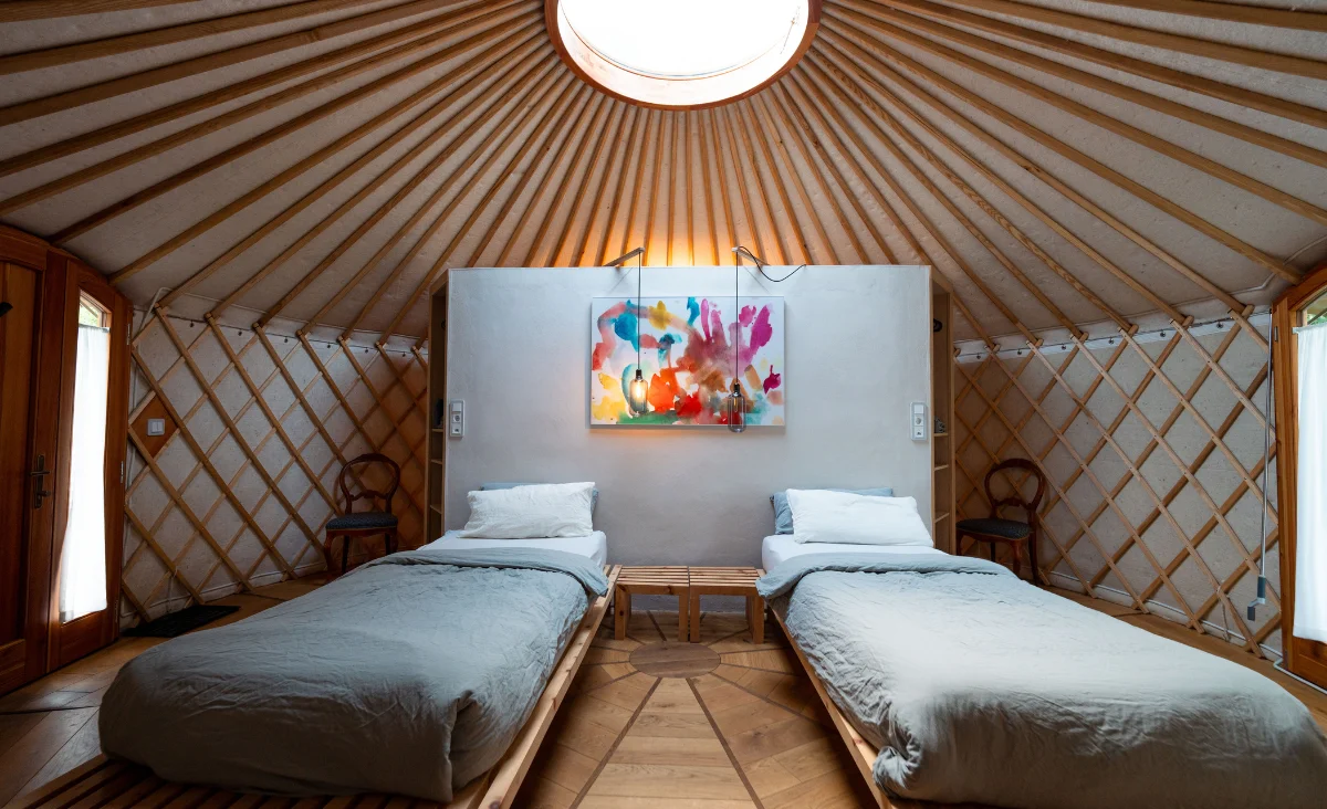 Innenraum eines runden Zeltes mit zwei Einzelbetten, einem kleinen Holztisch dazwischen und einem farbenfrohen Bild an der Wand unter einer runden Deckenöffnung