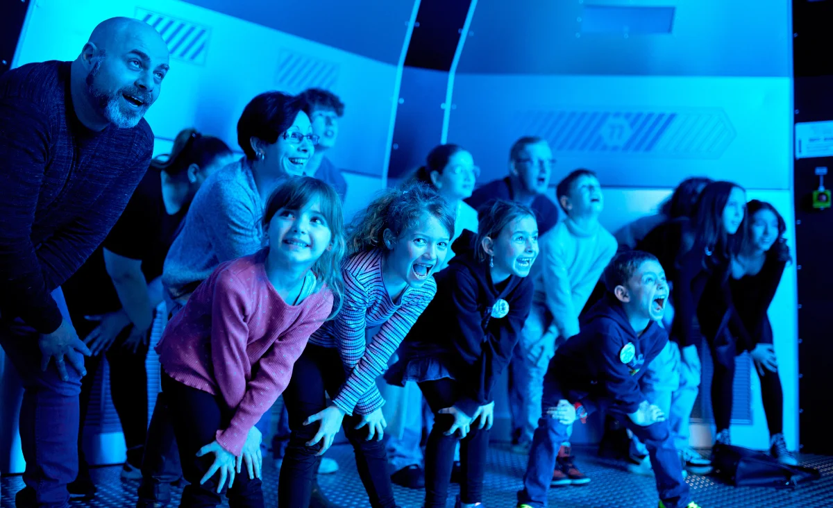 Gruppe von Kindern und Erwachsenen in gebückter Haltung mit aufgeregten Gesichtsausdrücken vor blau beleuchtetem Hintergrund