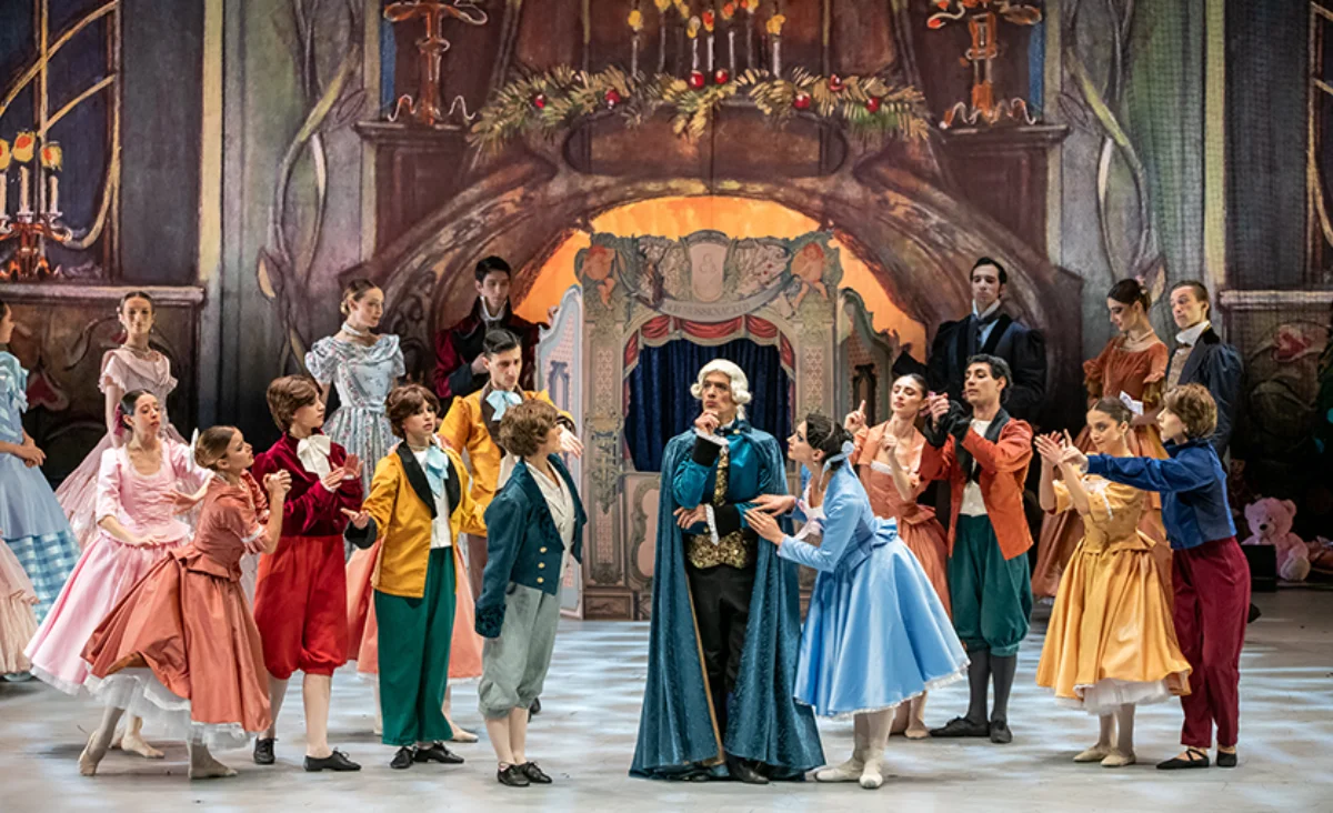 Mehrere Balletttänzer in farbenfrohen Kostümen stehen auf einer Bühne vor einem großen Kamin mit Kerzen und festlicher Dekoration, Szene aus dem Ballett "Der Nussknacker" des Grand Classic Ballet Berlin